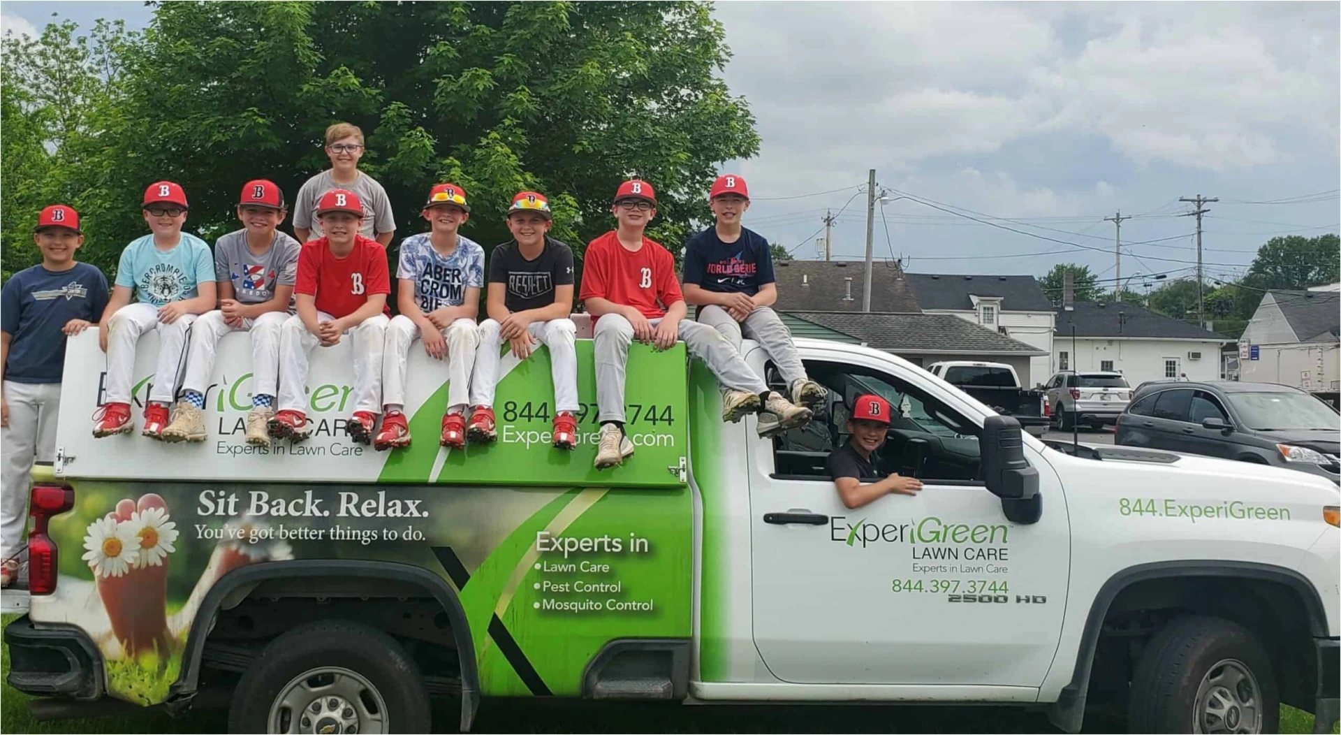 Group of young baseball players wearing red 'B' caps sitting on and around a white and green 'ExperiGreen Lawn Care' truck with a backdrop of buildings and cars under a cloudy sky.